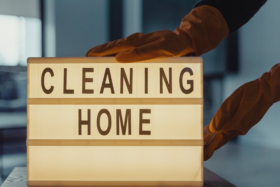A lightbox sign on a table displaying the words 'CLEANING HOME' in bold black letters on a white background. The sign appears to be illuminated, providing a warm glow. Behind the sign, there are two h